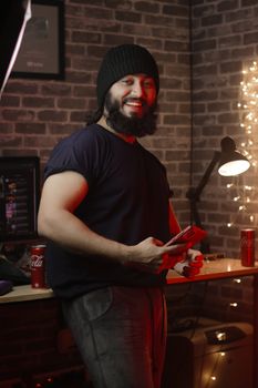 Smiling man with a beard holds a smartphone in a cozy indoor setting with warm lighting.