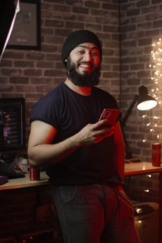 Bearded man in a casual setting using a smartphone with a smile.