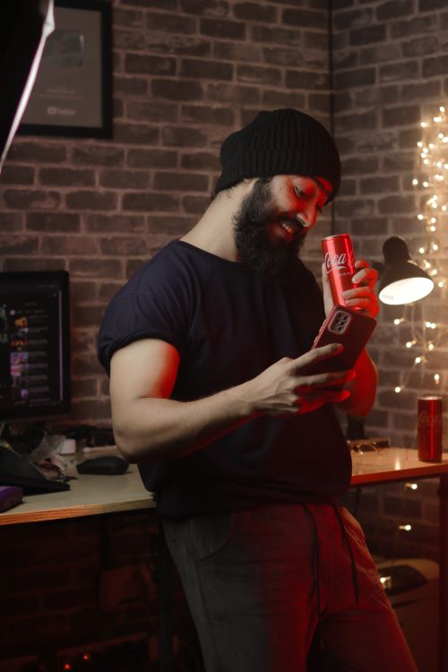 Free Young man with a beard holding a drink and smartphone indoors with a cozy ambiance and brick wall backdrop. Stock Photo