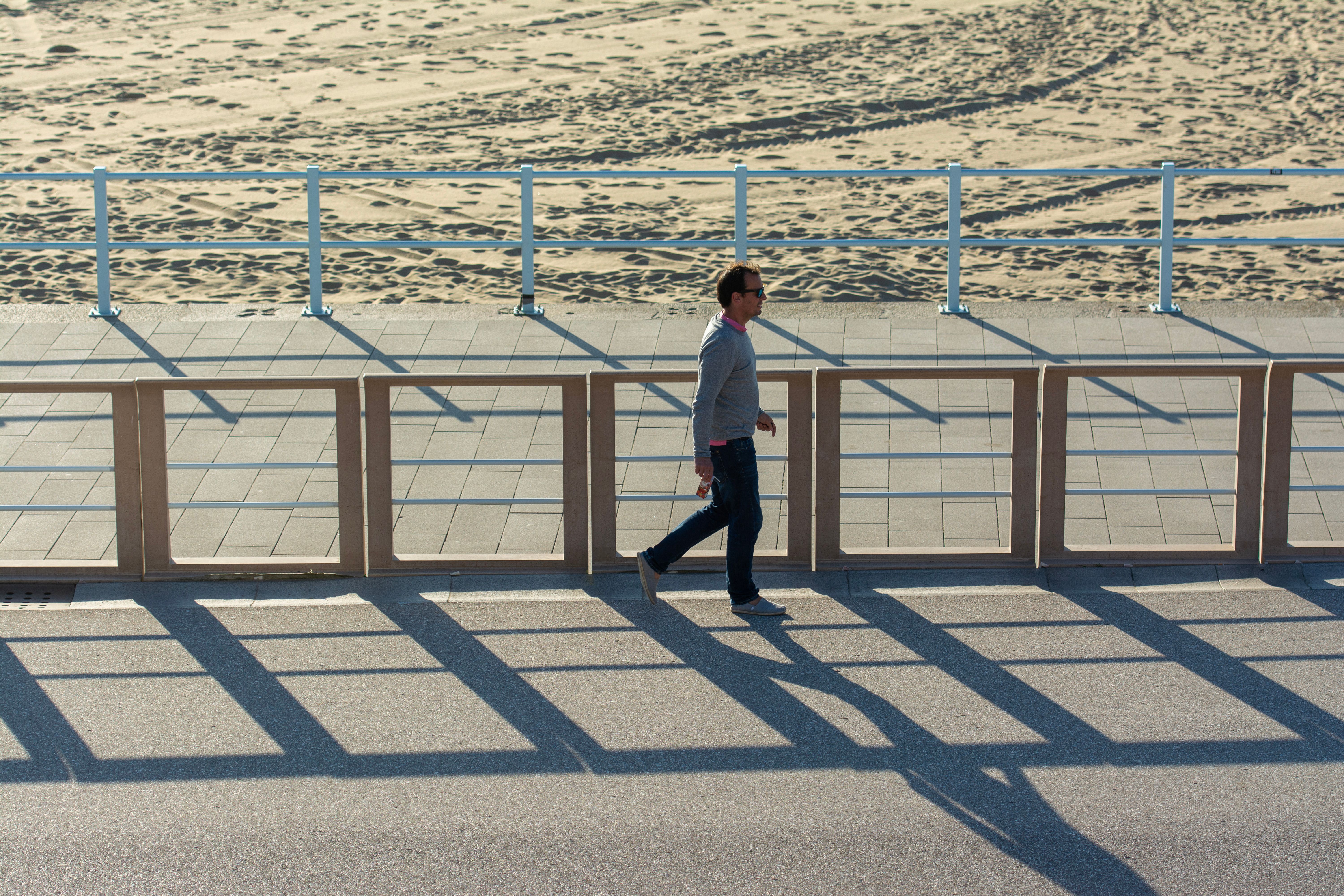 A solitary man walks on a sunny promenade casting shadows with a sandy beach backdrop. — Trójmiasto