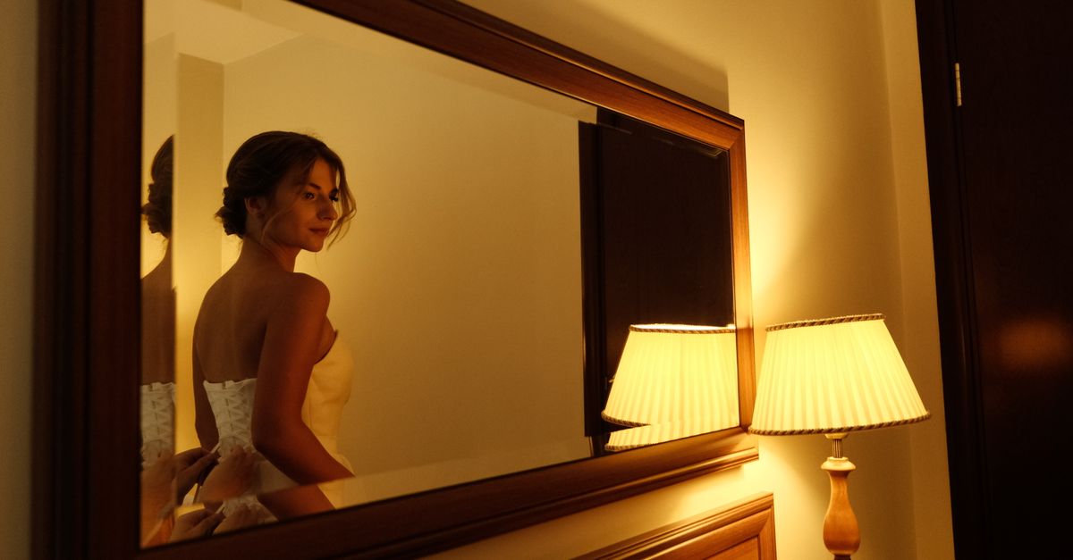 A woman in a white dress looks over her shoulder at her reflection in a warmly lit room.