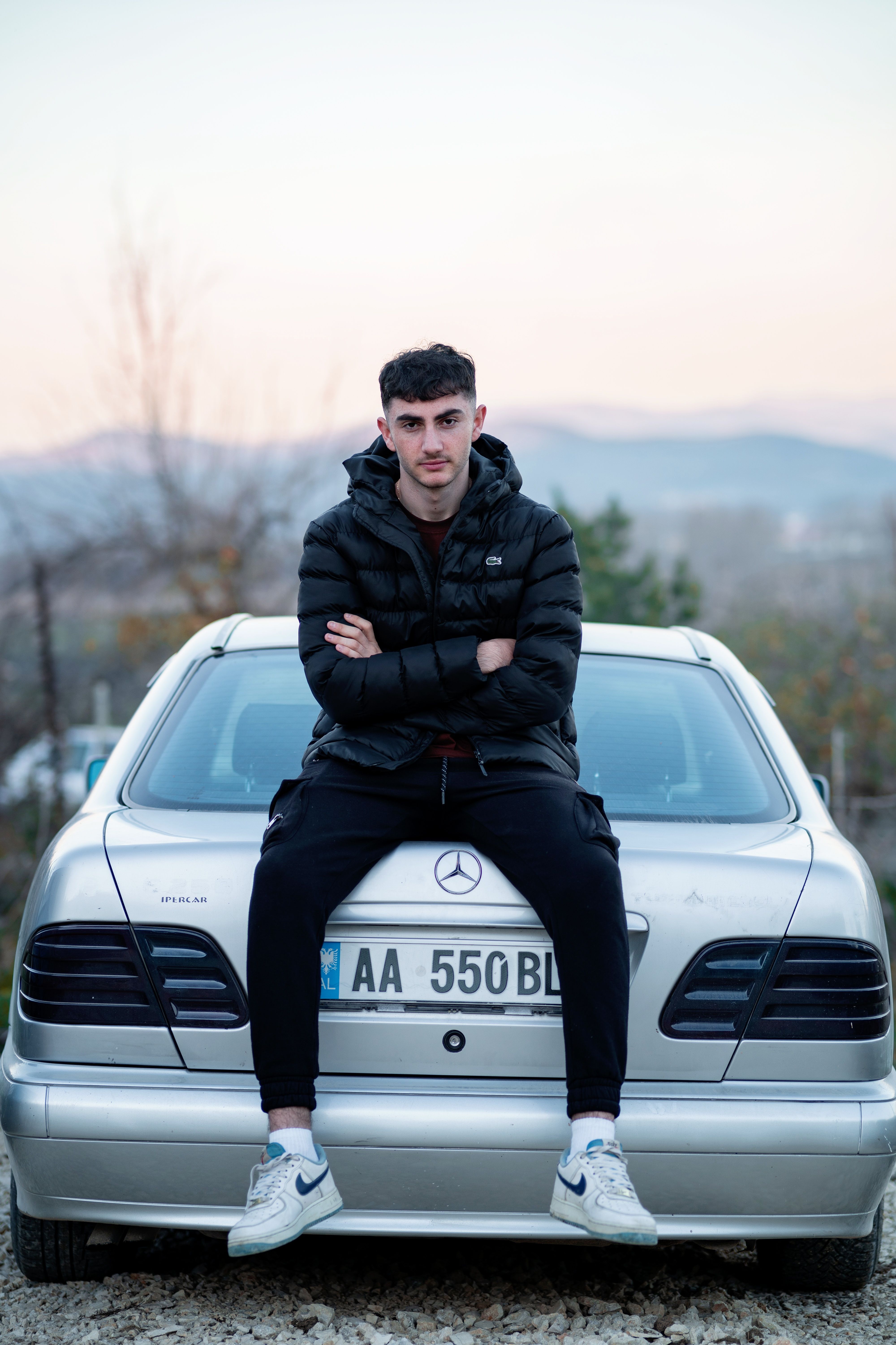 Young man in puffer jacket sitting on a Mercedes-Benz, arms crossed, during sunset outdoor scene.
