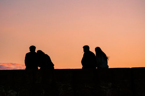 Free Silhouetted group of people sitting on a ledge against a vibrant sunset sky. Stock Photo