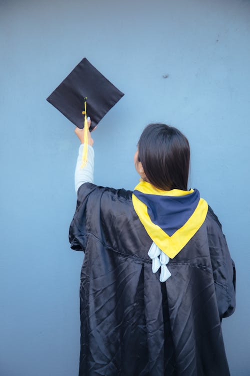 Back View of a Woman Wearing Black Graduation Gown while Holding a Square Graduation Cap