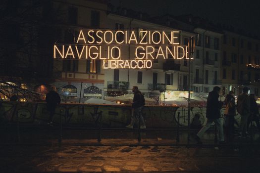 Street scene in Milan featuring people and neon sign at night.
