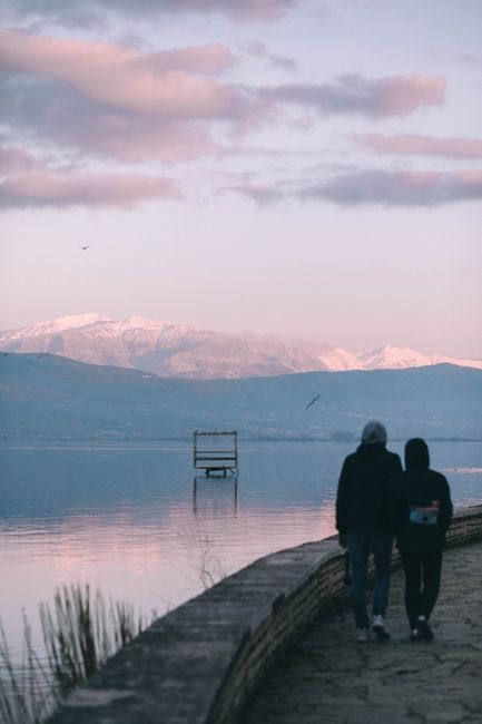 Couple strolling by a serene lake at sunset with mountains in the background in Ioannina, Greece.