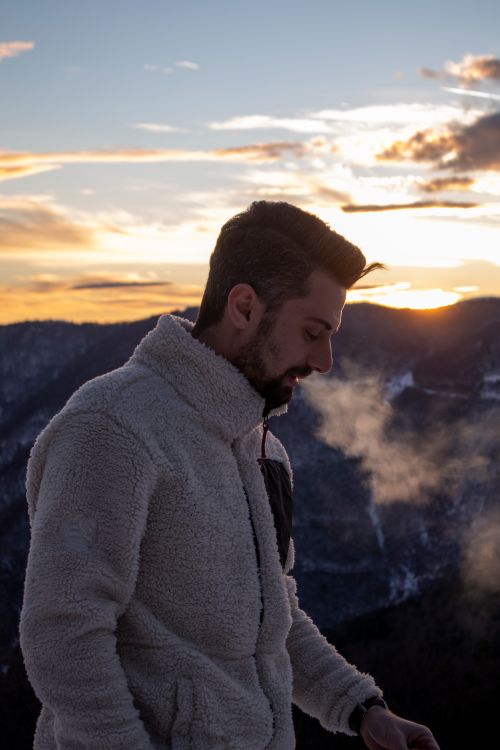 Free A man in warm clothing stands against a mountain backdrop at sunset, exhaling visible breath. Stock Photo
