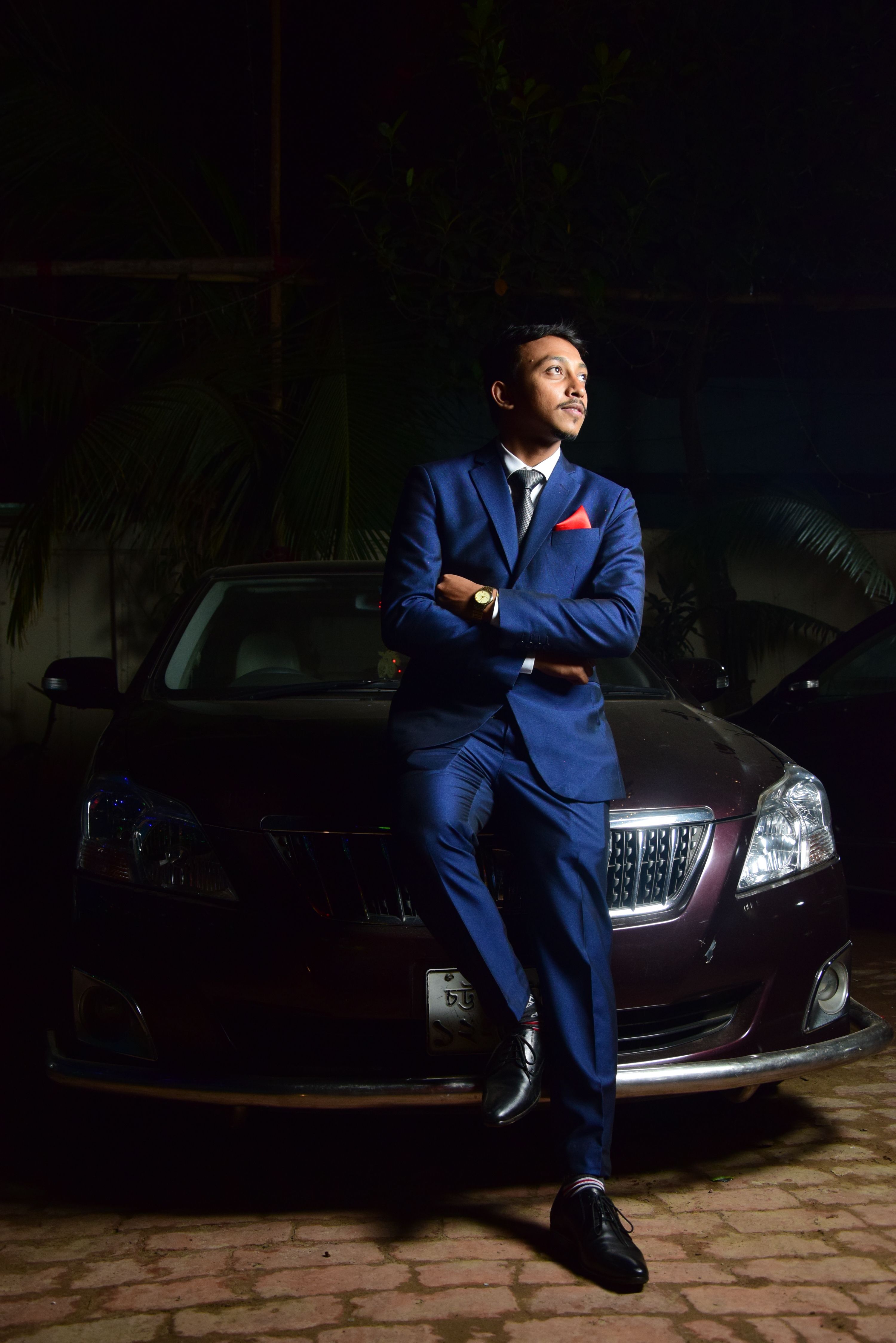 A fashionable man in a blue suit leans against a car, exuding confidence.