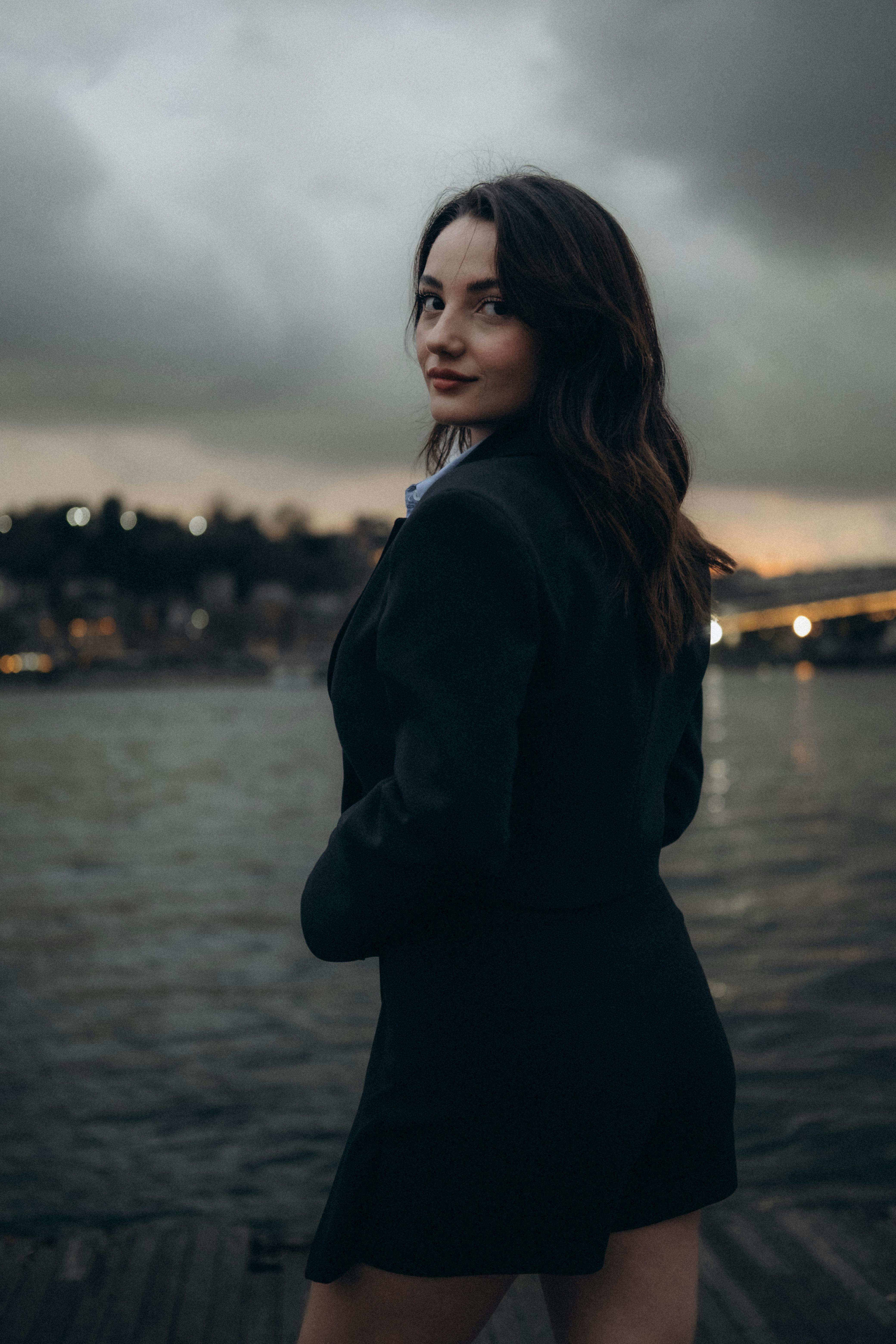 A woman in a black blazer and mini skirt poses by a river in Turkey during twilight, exuding elegance.
