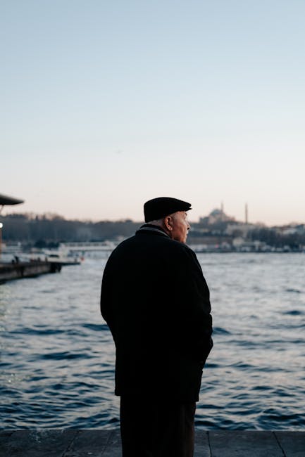 An elderly man stands by the river at dusk, with the city skyline in the background.