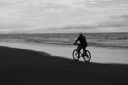 Free Black and white image of a cyclist on an overcast beach, capturing solitude. Stock Photo