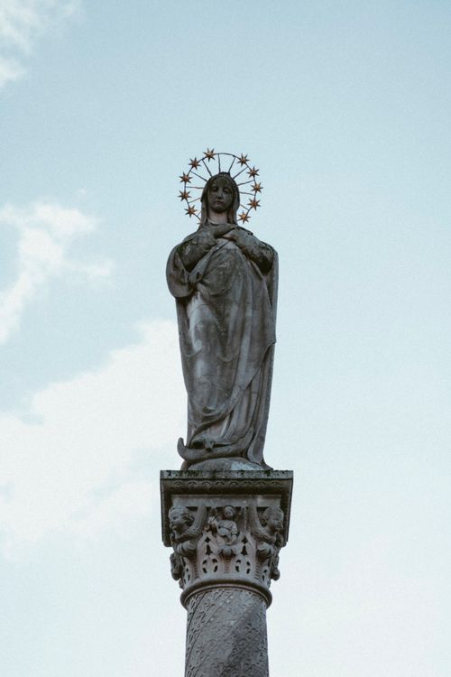 Free A sculpted figure of the Virgin Mary with a halo, standing atop a decorated column against a clear sky. Stock Photo