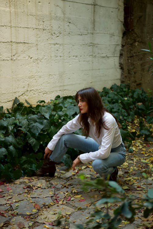 A Woman in White Long Sleeves and Denim Jeans Posing Beside Green Plants