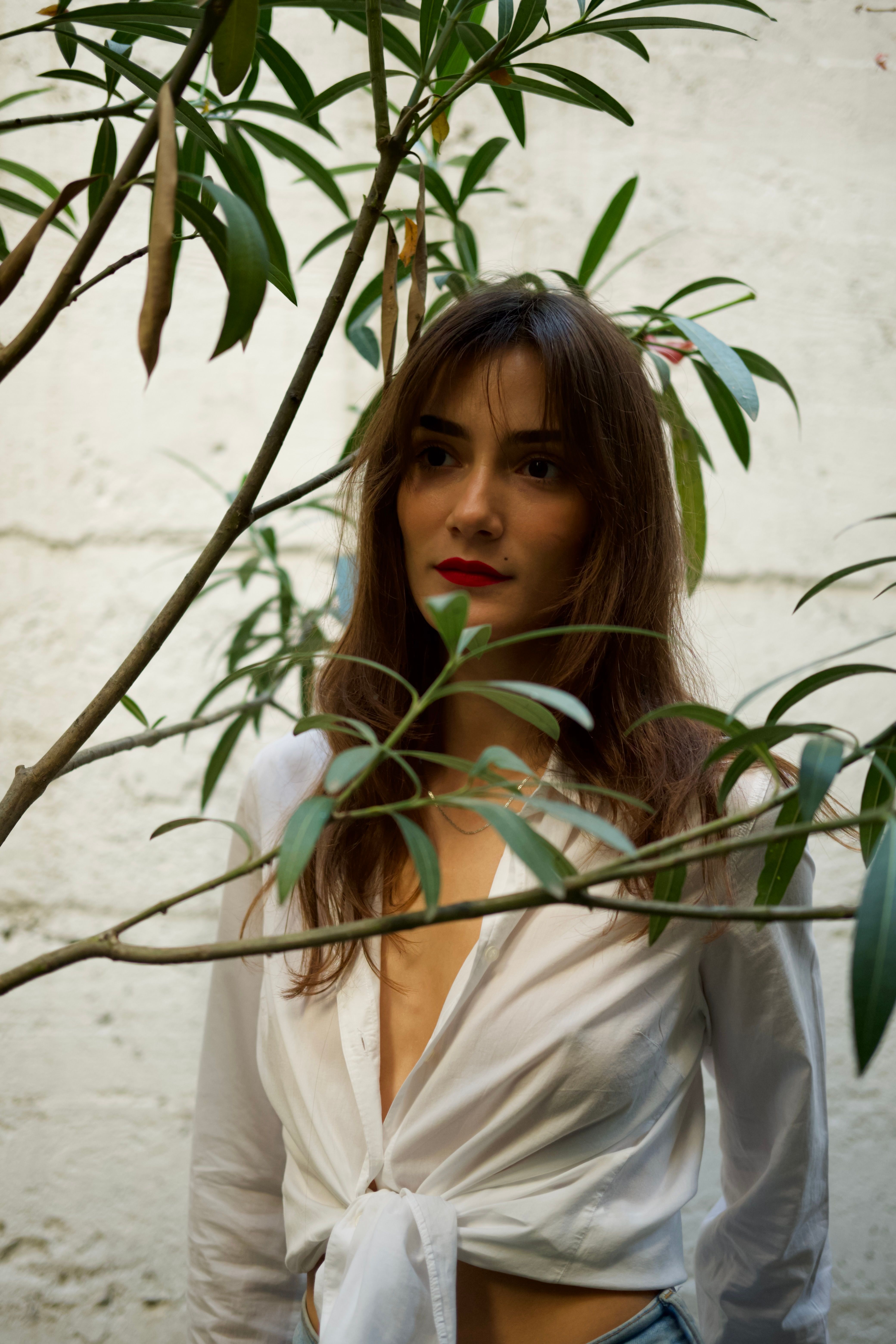 A woman with red lipstick stands outside in a white blouse by lush green leaves.