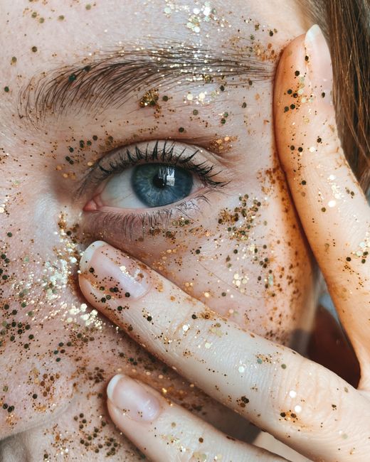 Dramatic close-up of a person's eye surrounded by glitter, with a hand gently touching the face.
