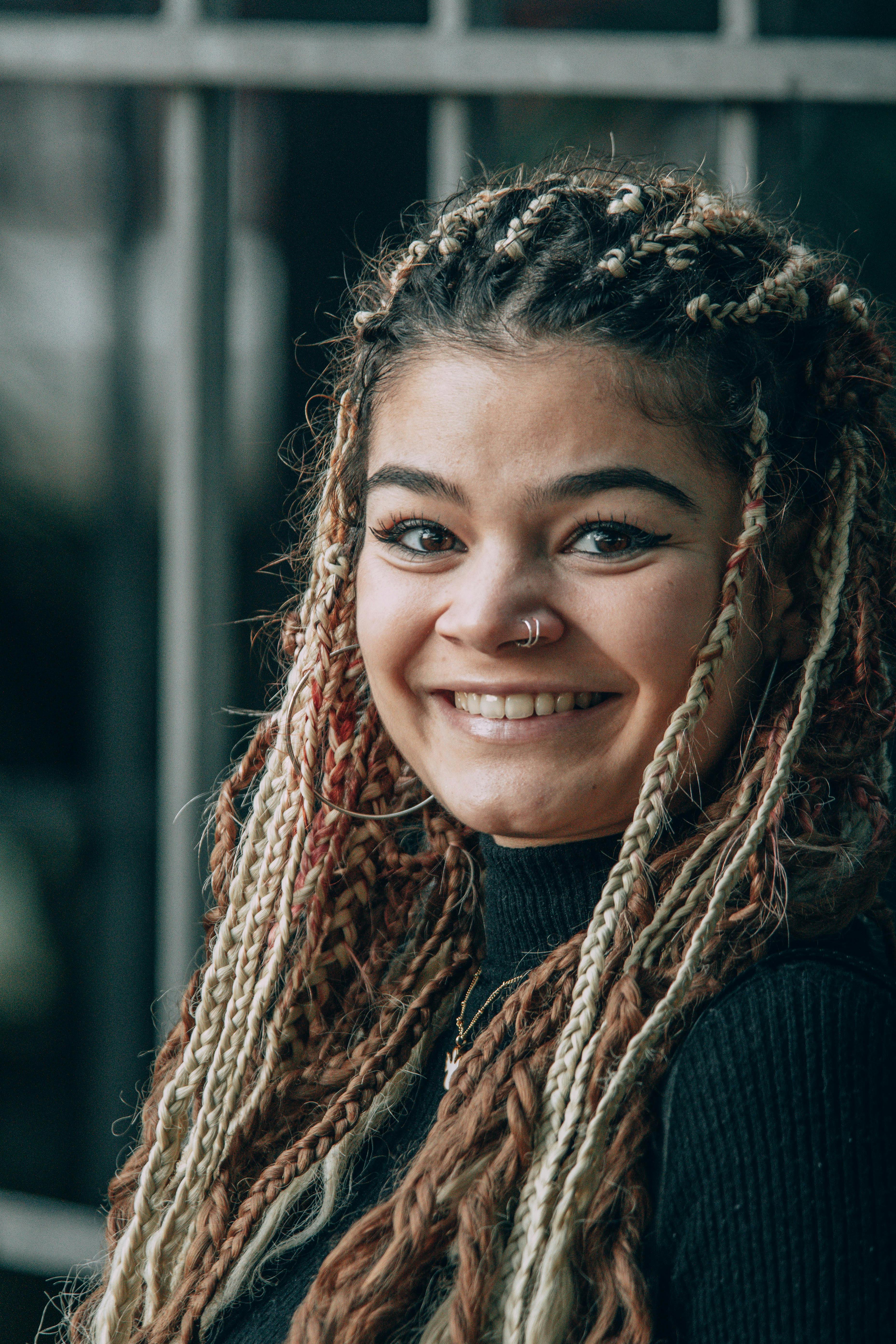 A young woman with braided hair and a nose piercing smiles warmly in a portrait.