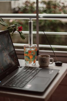 A serene home office setup with a laptop, coffee mug, plant, and incense by the window.