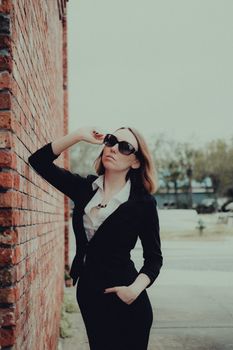Elegant businesswoman posing against a brick wall in sunglasses, exuding confidence in a chic outdoor setting.