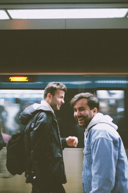 Two men smiling on a subway platform as a train rushes by in an urban setting.