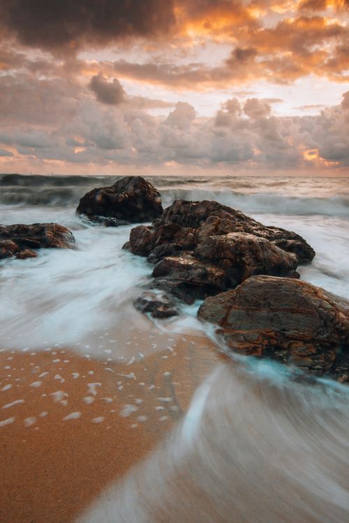 Free Capture of waves crashing against rocks on a beach during a dramatic sunset with vibrant clouds. Stock Photo