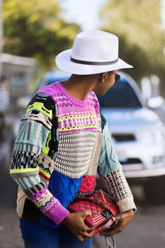 Stylish woman wearing a colorful sweater and hat, holding a red bag on a sunny day outdoors.