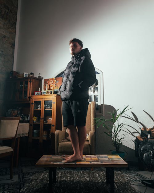 Photo of a Man Wearing a Winter Jacket in an Interior, and Standing on a Coffee Table