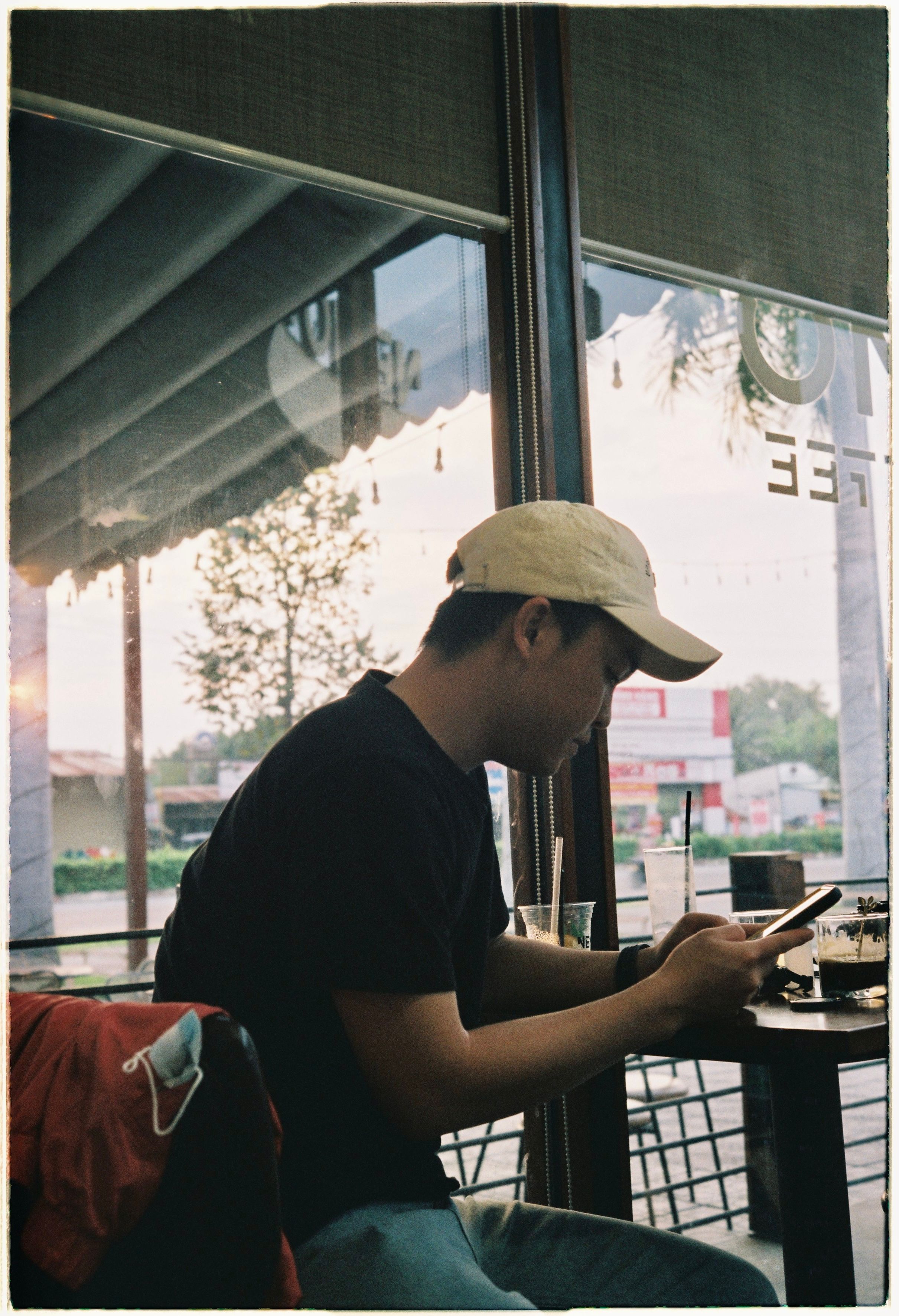Asian man wearing a yellow cap, sitting in café, focused on smartphone use by the window.