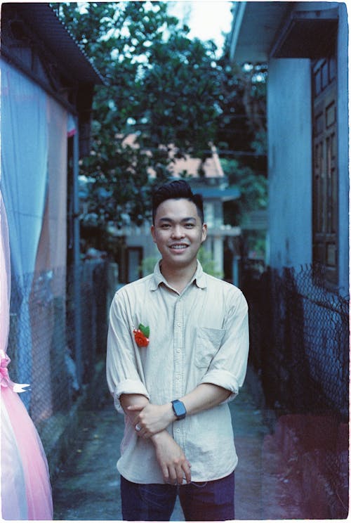 Photo of a Young Man with a Floral Brooch Standing by Village Houses 