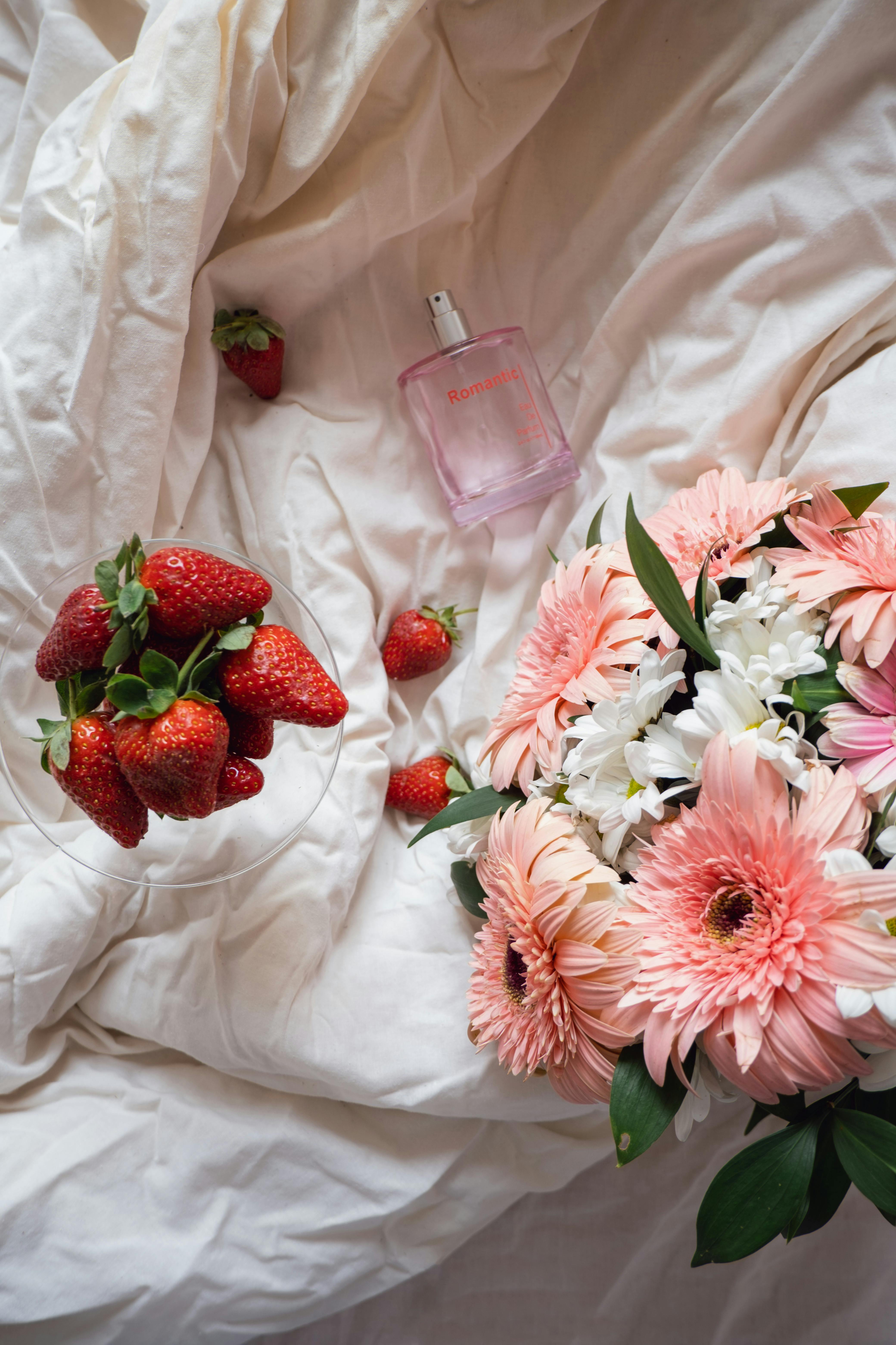 Flat lay of pink flowers, strawberries, and perfume on white linen for a romantic setting.