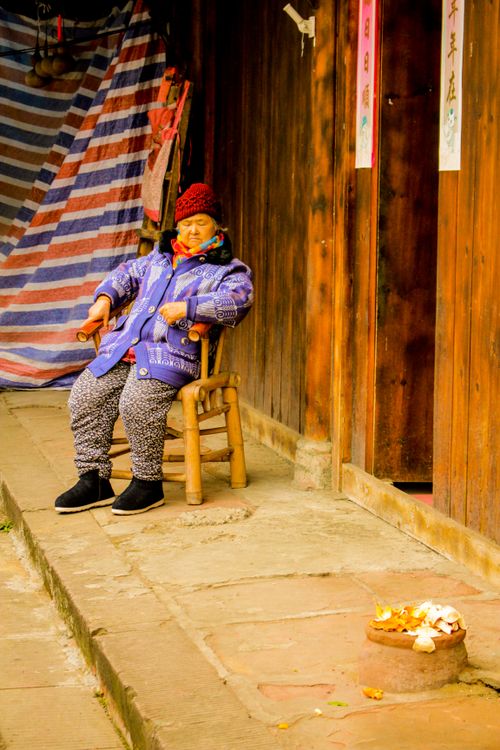 Free An elderly woman in a colorful jacket sits by a wooden village house, enjoying a peaceful moment. Stock Photo