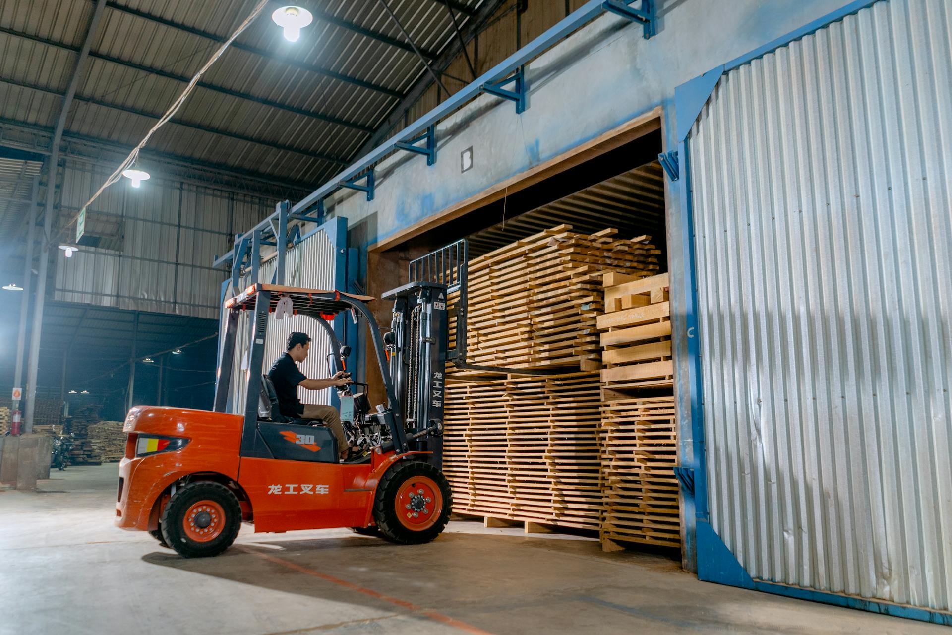 Man operating a forklift indoors loading wooden pallets in a warehouse.