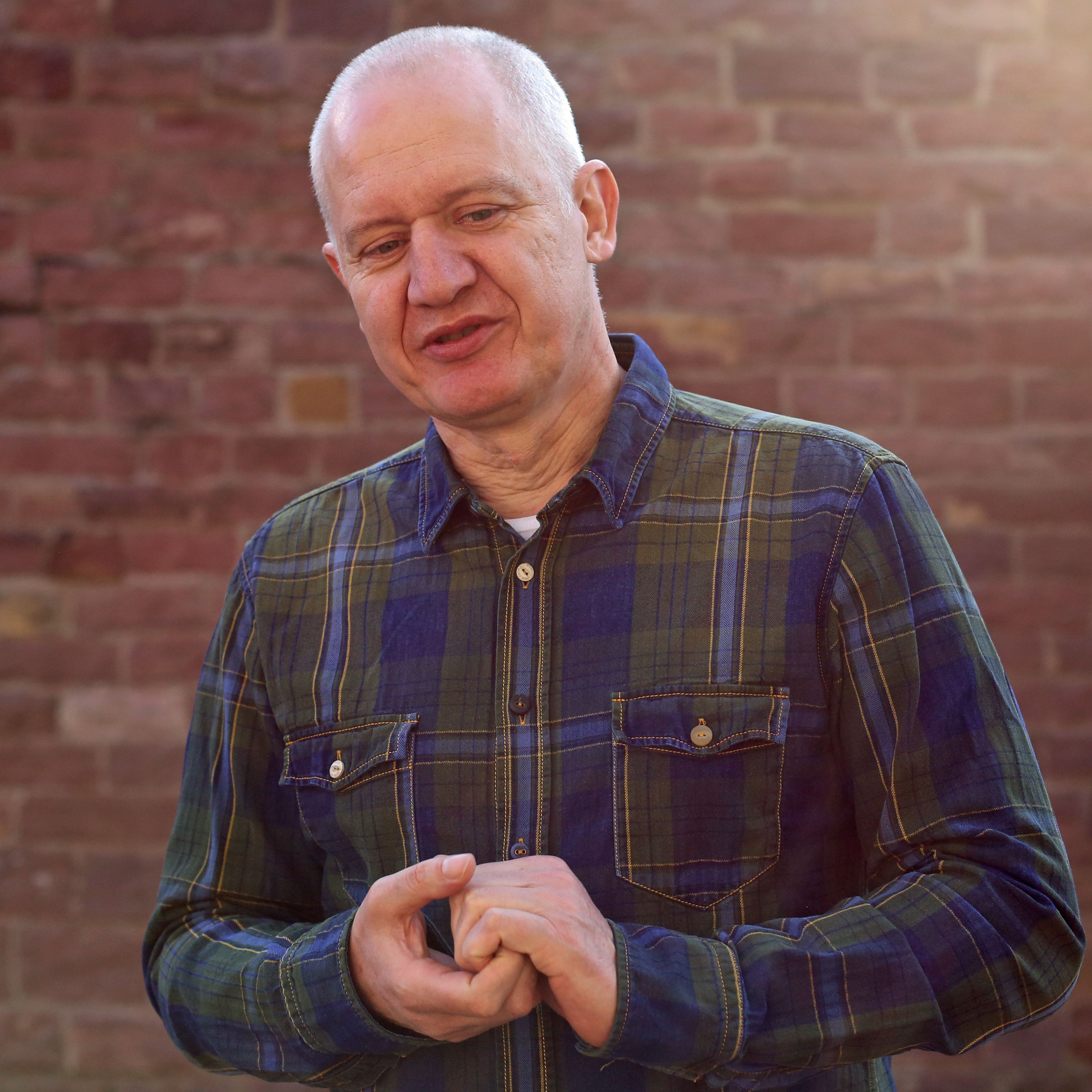 Close-up portrait of an elderly man wearing a checkered shirt against a brick wall.