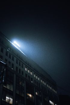 Dramatic night view of an illuminated office building under rain, showcasing urban architecture.
