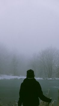 A solitary figure stands by a foggy lake surrounded by winter snow, evoking mystery.
