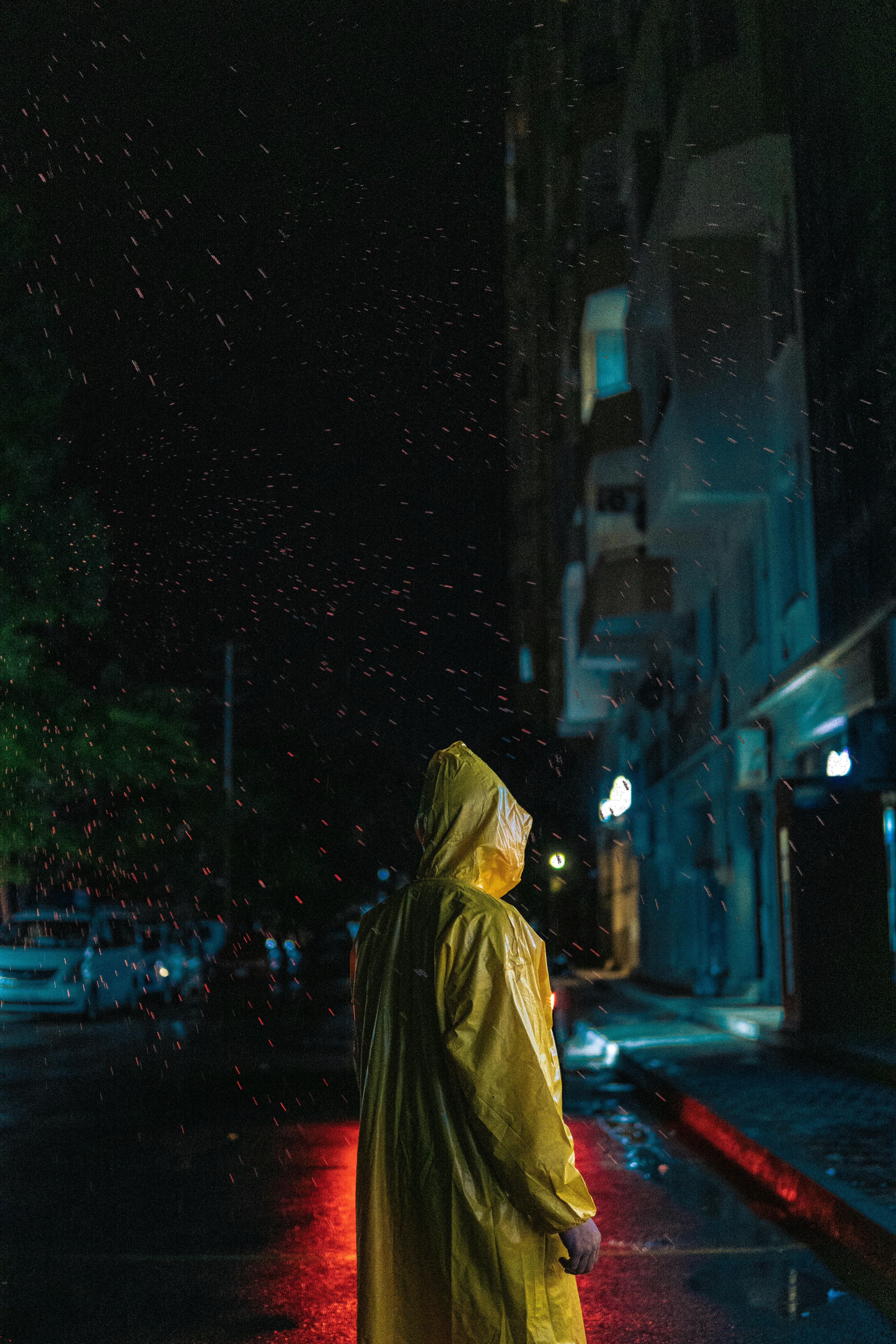A person in a yellow raincoat stands on a rainy street at night, with city lights reflecting on wet pavement.