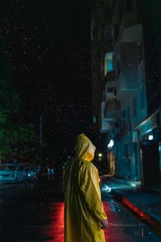 A person in a yellow raincoat stands on a rainy street at night, with city lights reflecting on wet pavement.