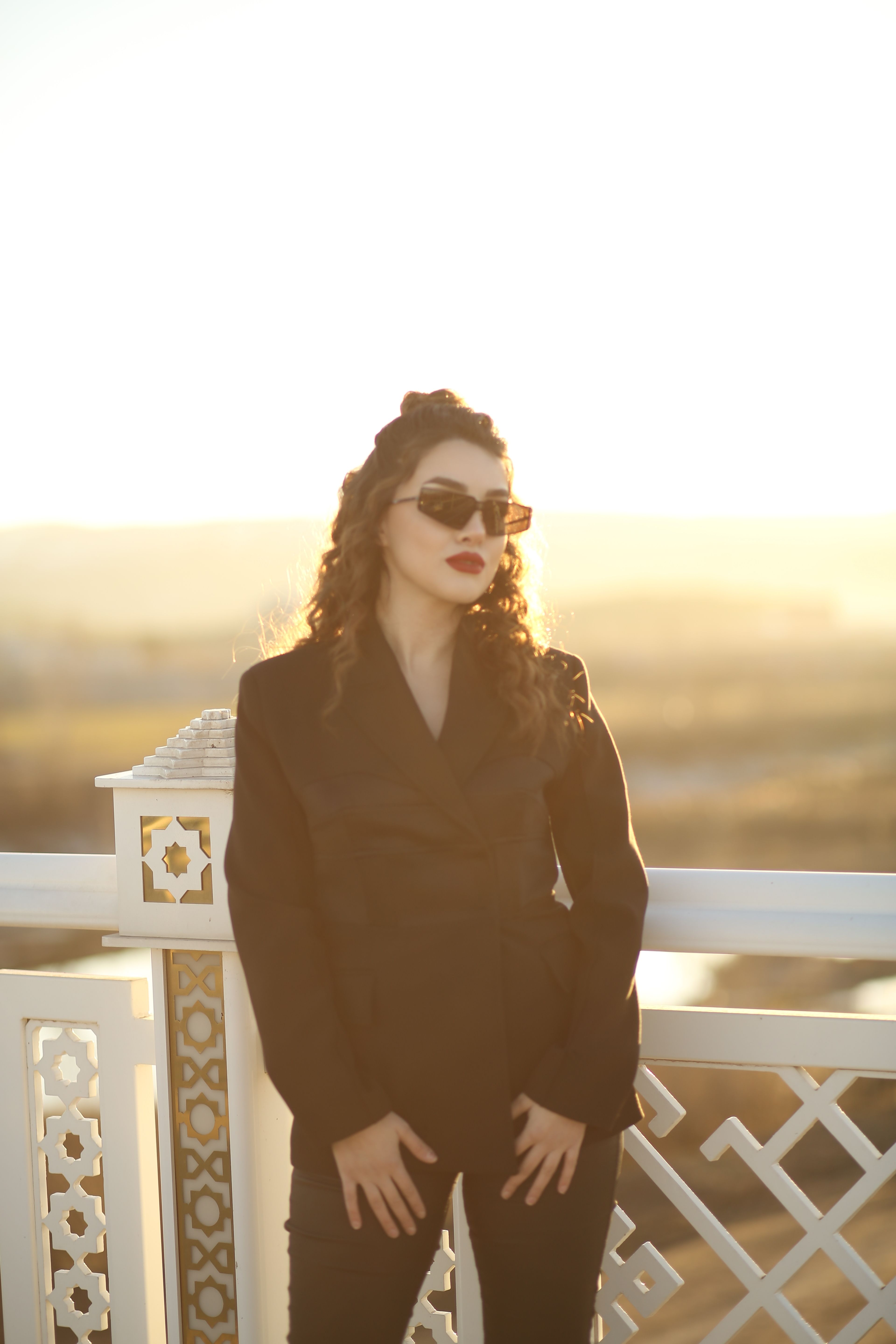 Stylish woman in a black suit and sunglasses poses elegantly outdoors during sunset.