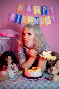 Woman enjoying birthday cake surrounded by dolls under a colorful banner.