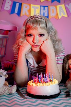 A young woman wearing a tiara ponders at her birthday party with a cake full of candles.