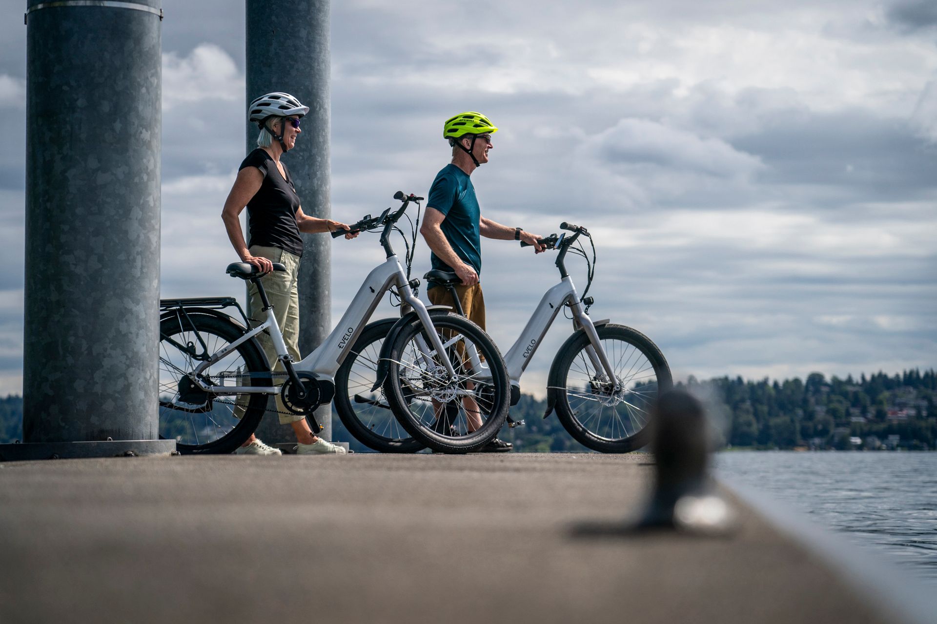 A couple on electric bikes pause on a pier in Seattle, overlooking a serene water scene.