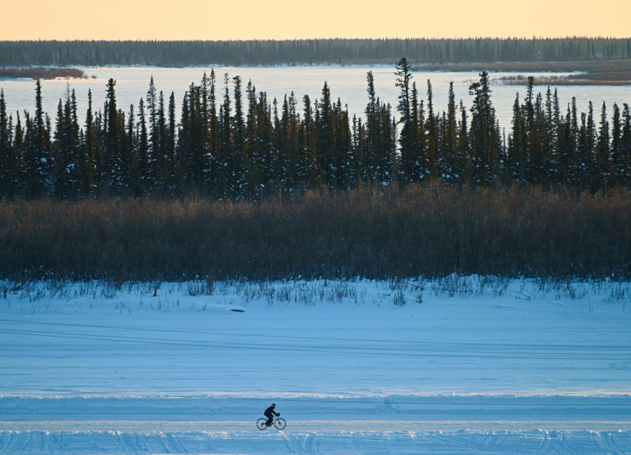 A person biking across a snowy and frozen landscape with trees and a lake, captured near Inuvik, NT, Canada.