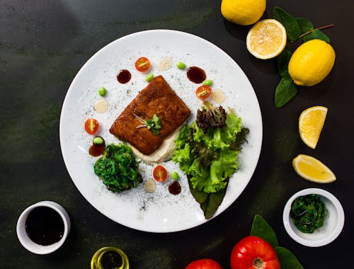 Free Top view of delicious fried fish served in white plate with fresh salad and spinach and placed on black table table with lemons tomatoes and sauces Stock Photo