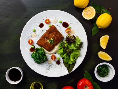 Top view of delicious fried fish served in white plate with fresh salad and spinach and placed on black table table with lemons tomatoes and sauces