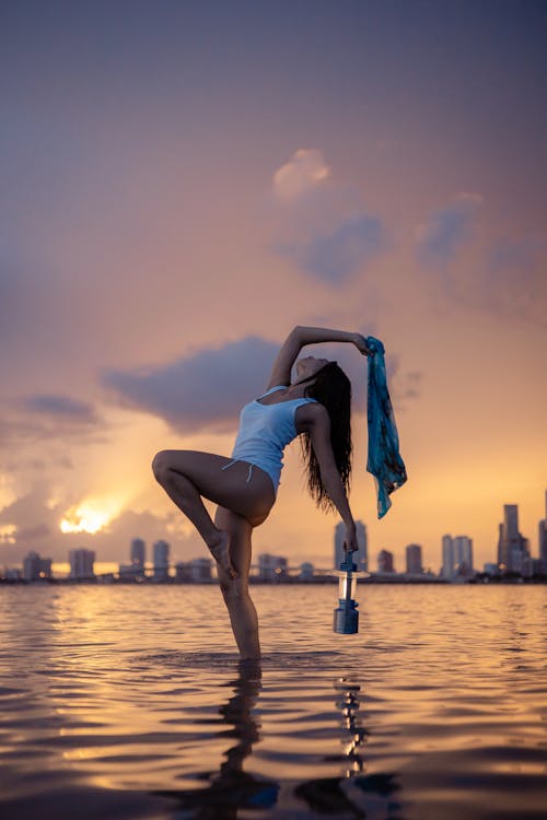 Woman Posing with Lamp in Water at Sunset with City behind