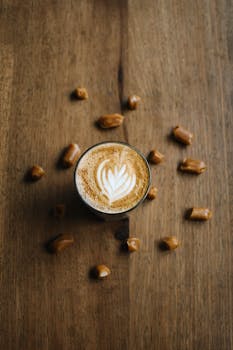 A latte with intricate latte art surrounded by caramel pieces on a wooden table.