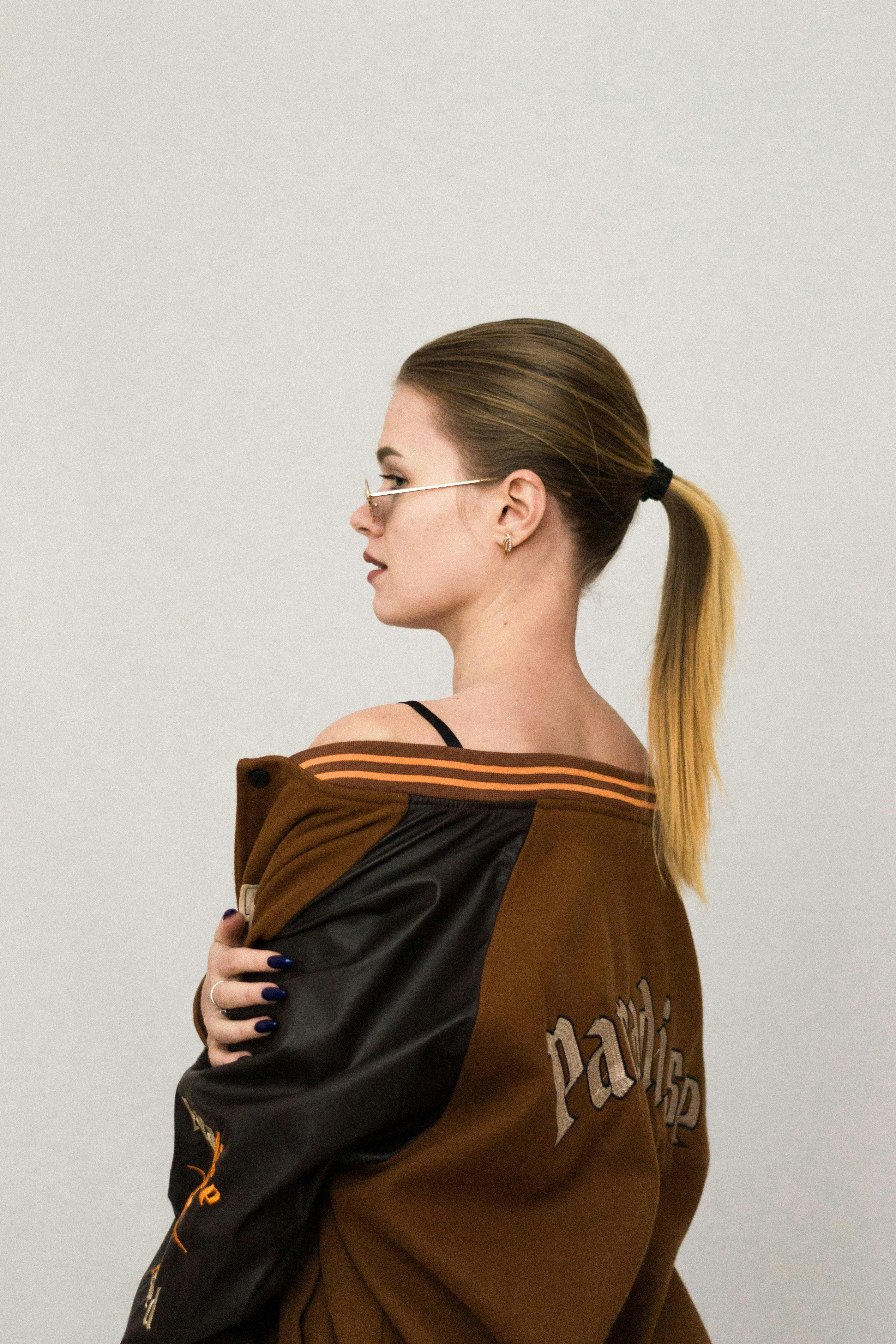 Side profile of a woman in a varsity jacket, sunglasses, and ponytail displaying modern style.