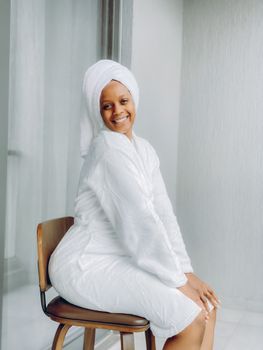 Cheerful woman in a white bathrobe sitting and smiling indoors with a towel wrap.