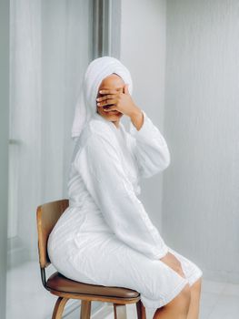 Woman in a bathrobe with towel-covered hair sits indoors, covering her face for a private moment.