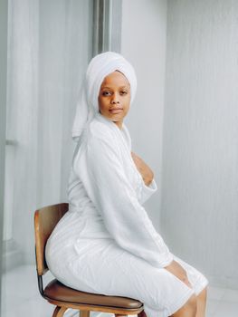 Woman in a white bathrobe sitting on a chair in a bright indoor setting, exuding calmness and relaxation.