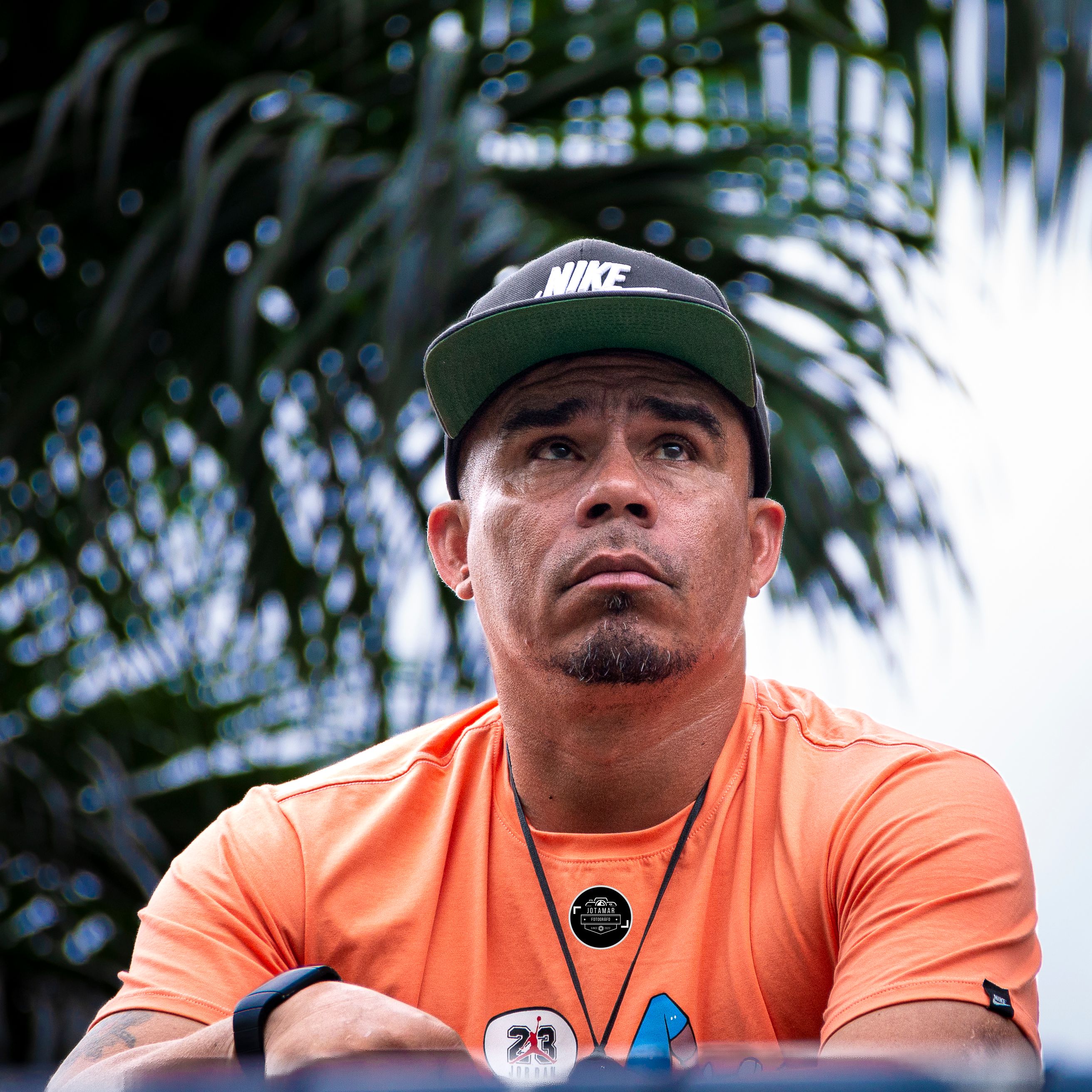 A man wearing a cap and t-shirt, looking pensive beneath palm trees.